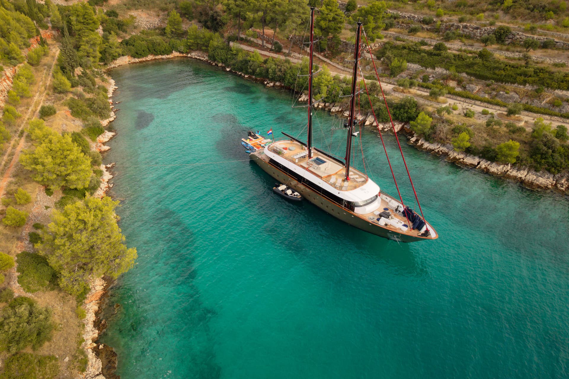 Large sailing yacht anchored in turquoise bay next to wooded, terraced coastline.