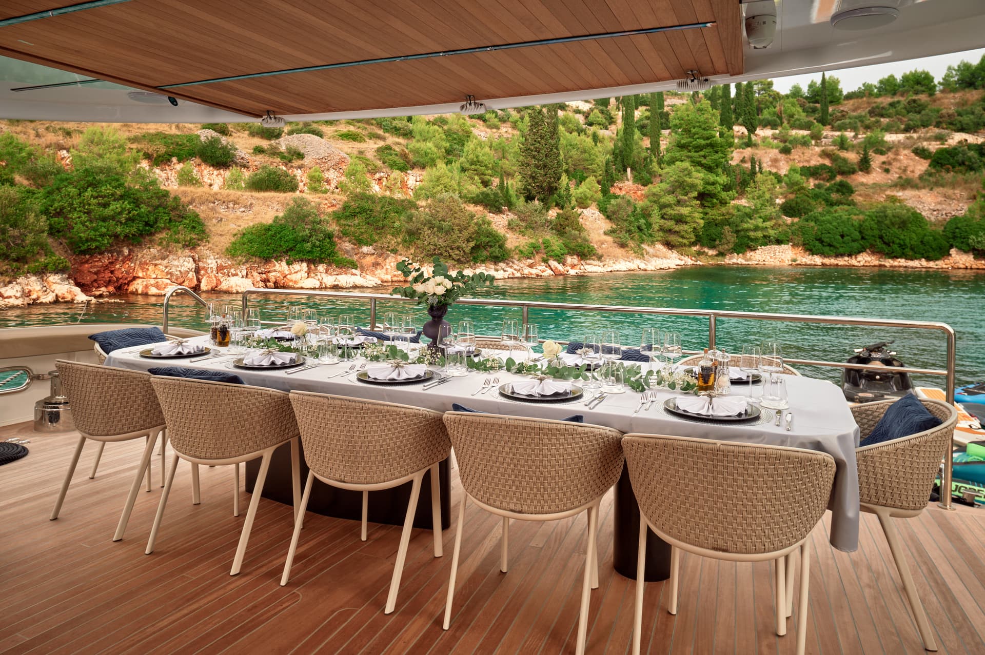 Anetta yacht main deck aft dining table set against a lush, green, rocky coastline.
