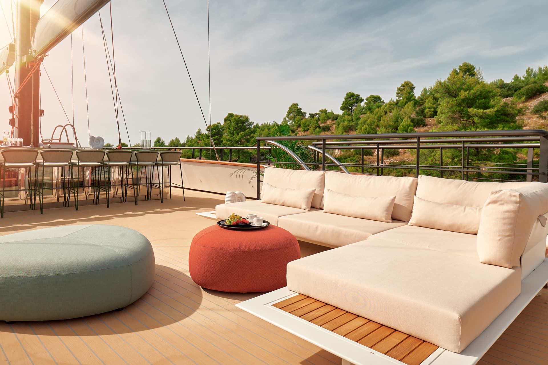 Sundeck lounge area with beige cushions, bar stools, and fruit tray overlooking a wooded hillside.