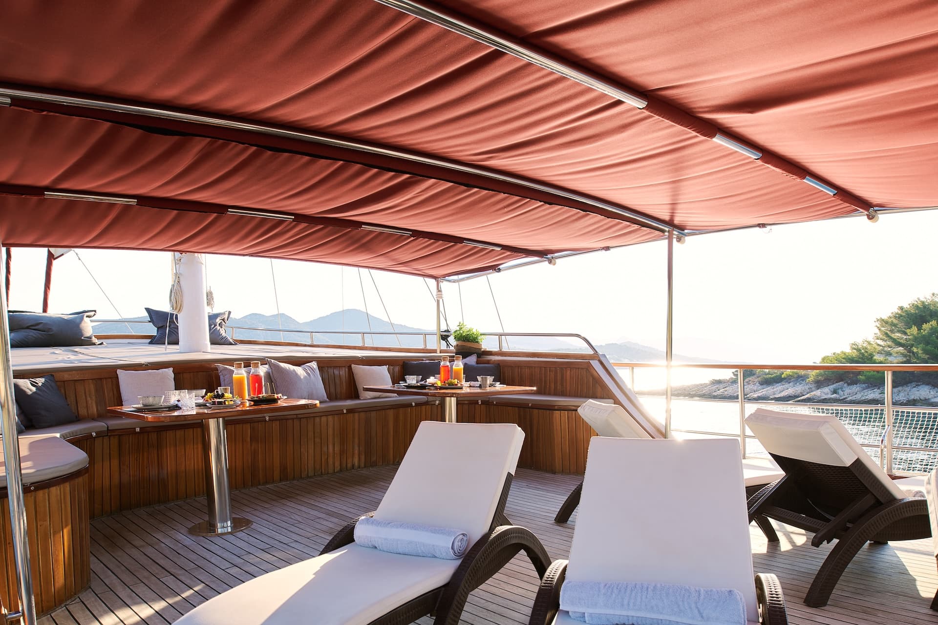 Sun deck with lounge chairs and breakfast table on a boat near a sunny coastline.