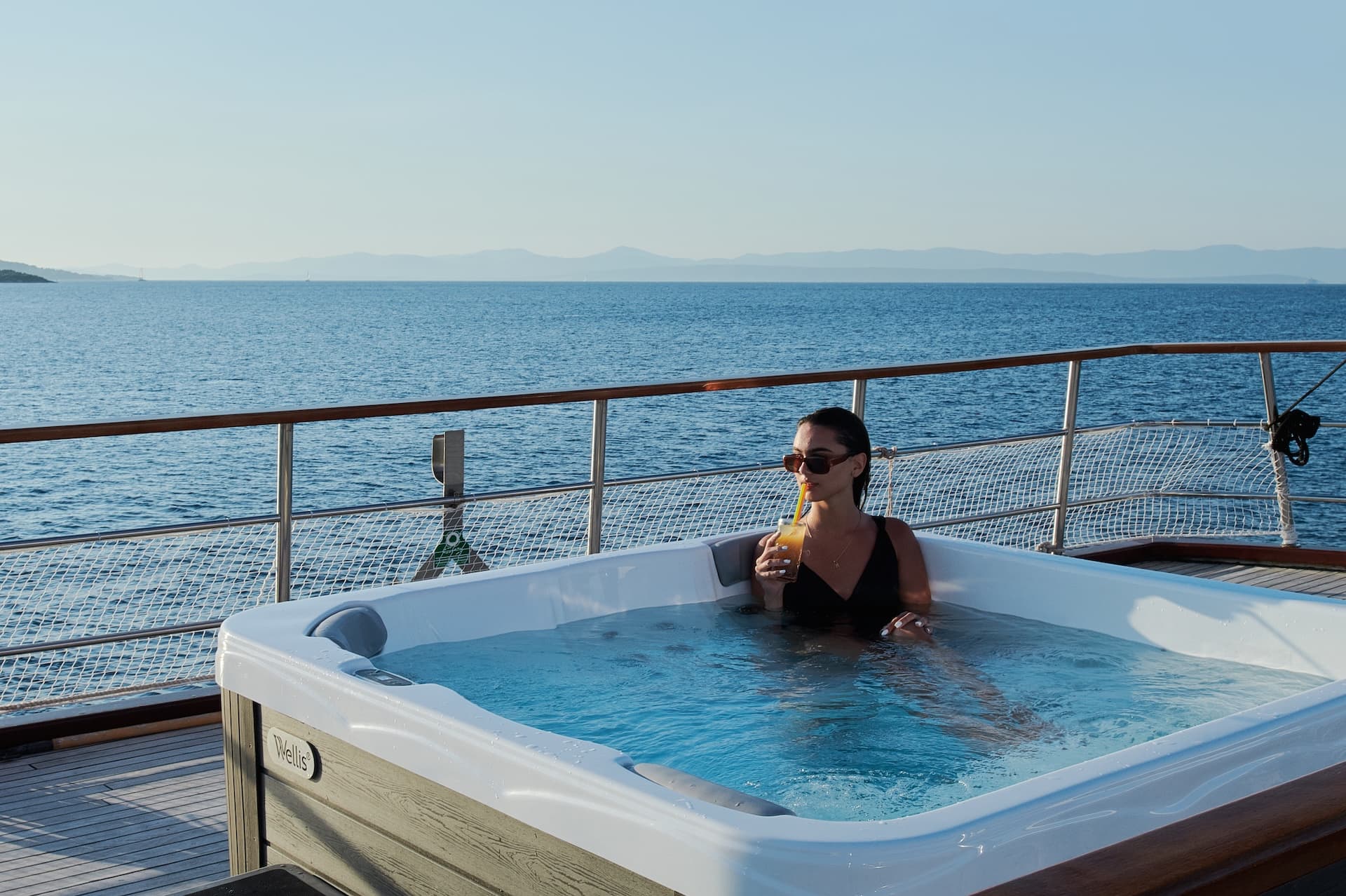 Woman relaxing in jacuzzi on deck overlooking calm blue sea and distant mountains
