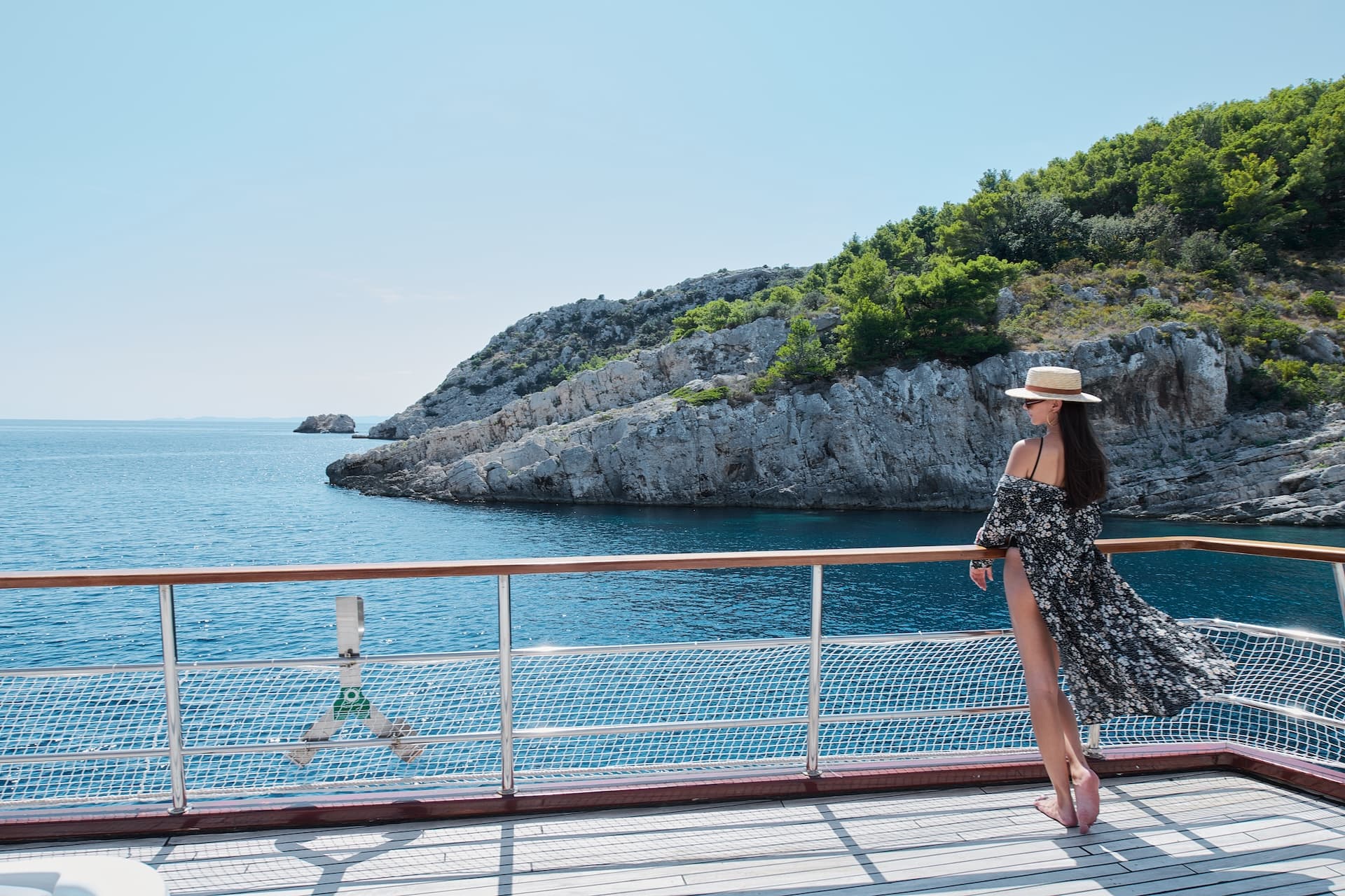 Woman on cruise deck looking at rocky, tree-covered coastline and blue sea.