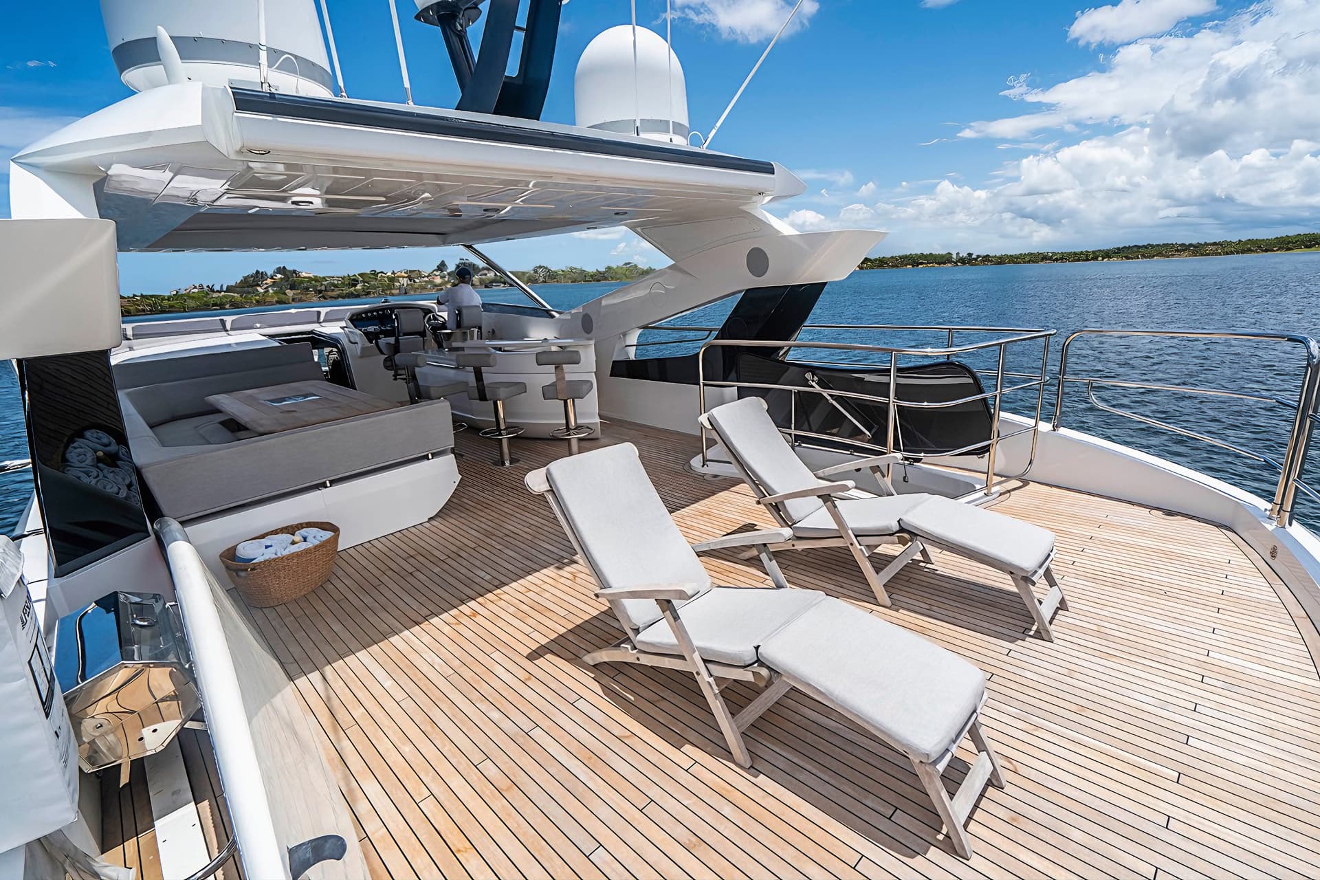 Luxury yacht deck with sun loungers, teak flooring, and coastal view under blue sky.