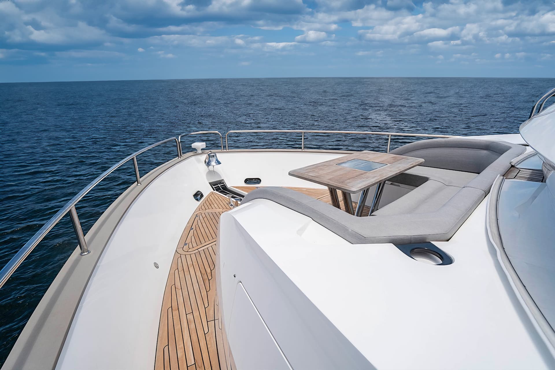 Bow seating area of a white yacht with teak decking on dark blue water under cloudy sky.