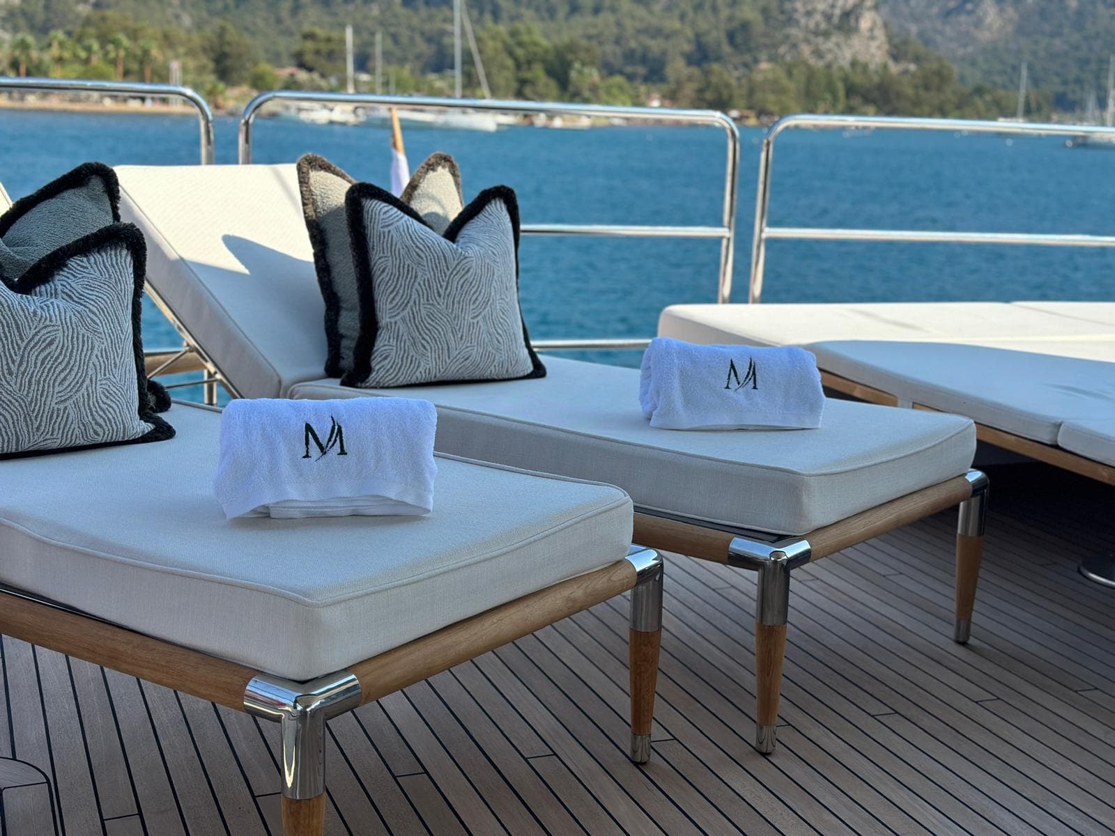 Lounge chairs with monogrammed towels on a yacht deck overlooking blue coastal waters and hills.