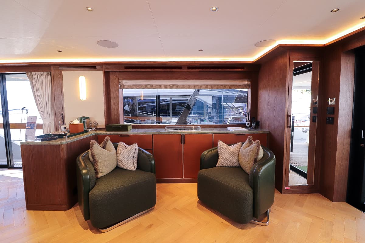 Saloon interior with wood paneling, two green armchairs, and view of a yacht through a window.