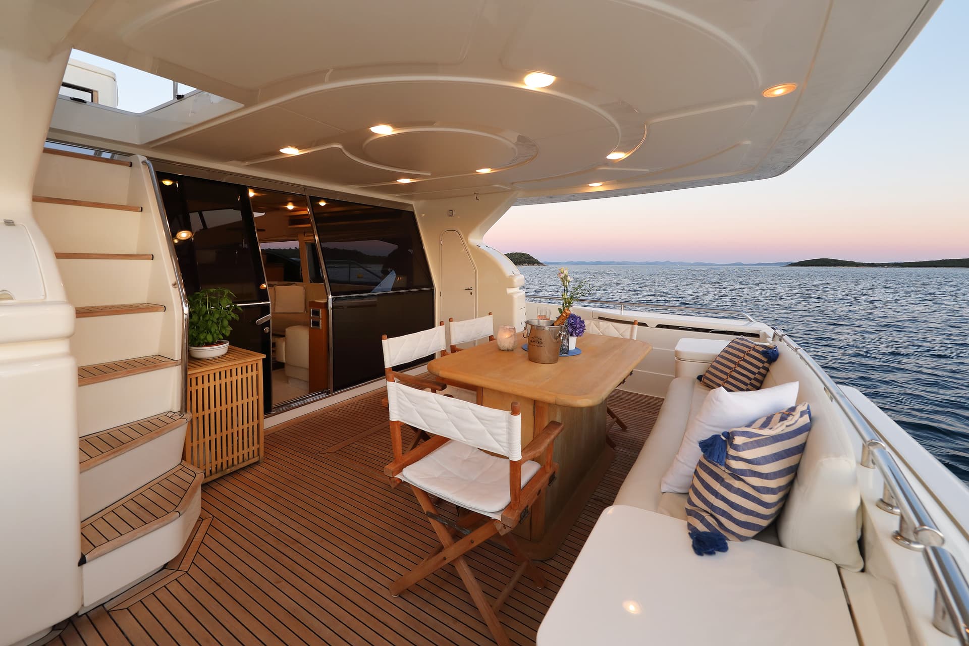 Yacht aft deck dining area with teak flooring overlooking the sea at sunset near islands.