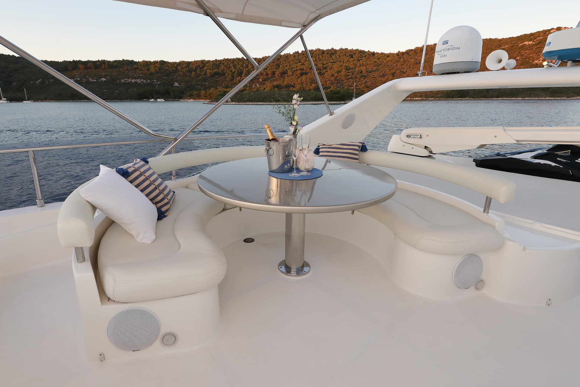 Flybridge seating area with champagne bucket and pillows overlooking a wooded coastline at sunset.