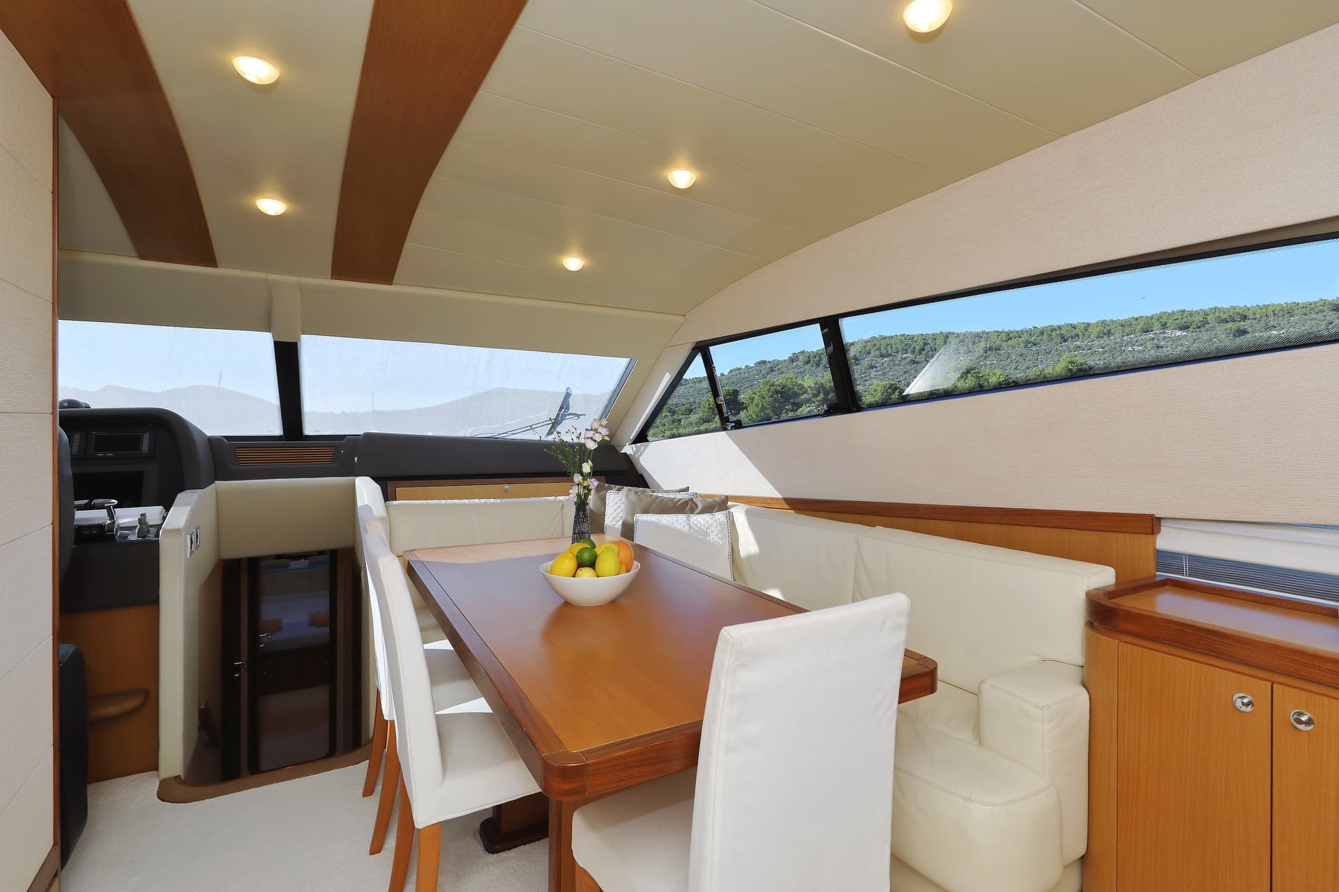 Yacht interior dining area with wood table, white seating, and view of green hills.