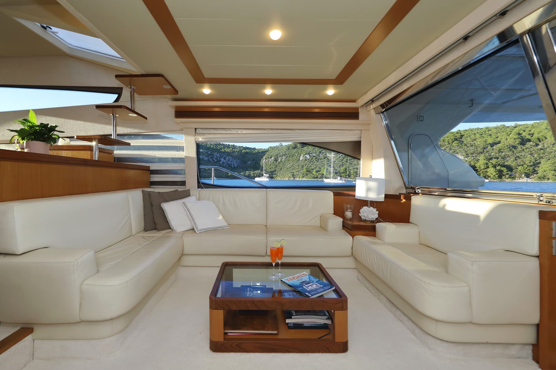 Luxury saloon interior of a yacht with white leather seating overlooking a wooded coastline and sailboats.