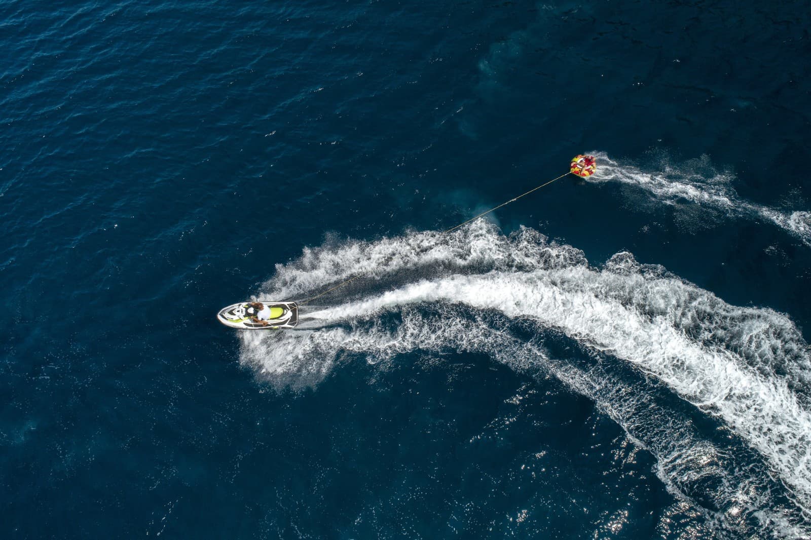 Jet ski towing an inflatable tube over deep blue coastal waters, aerial view