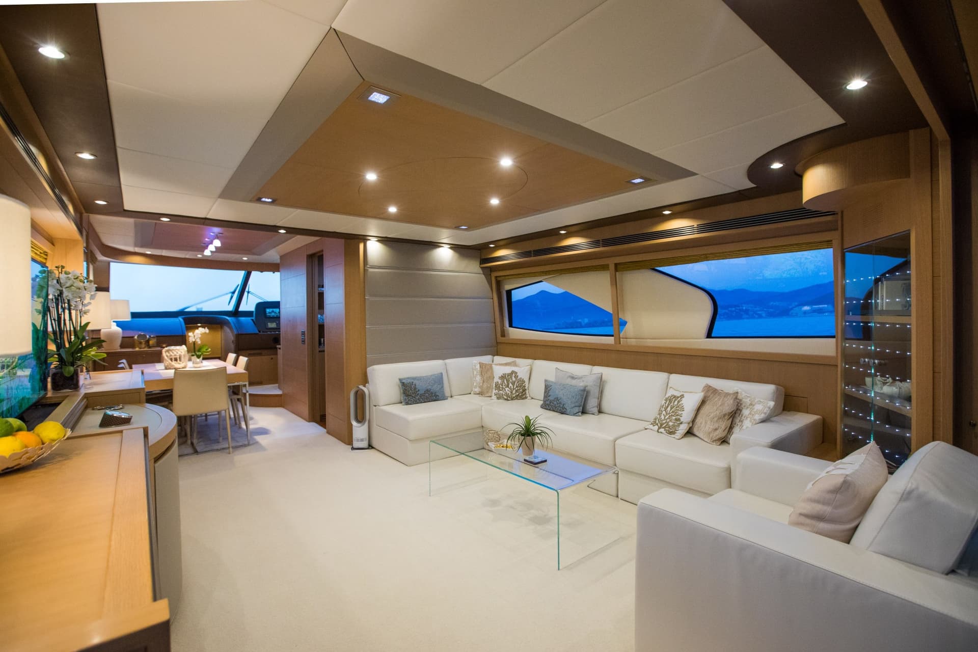 Luxury yacht interior with white sectional sofa, glass table, and view of blue water and mountains.
