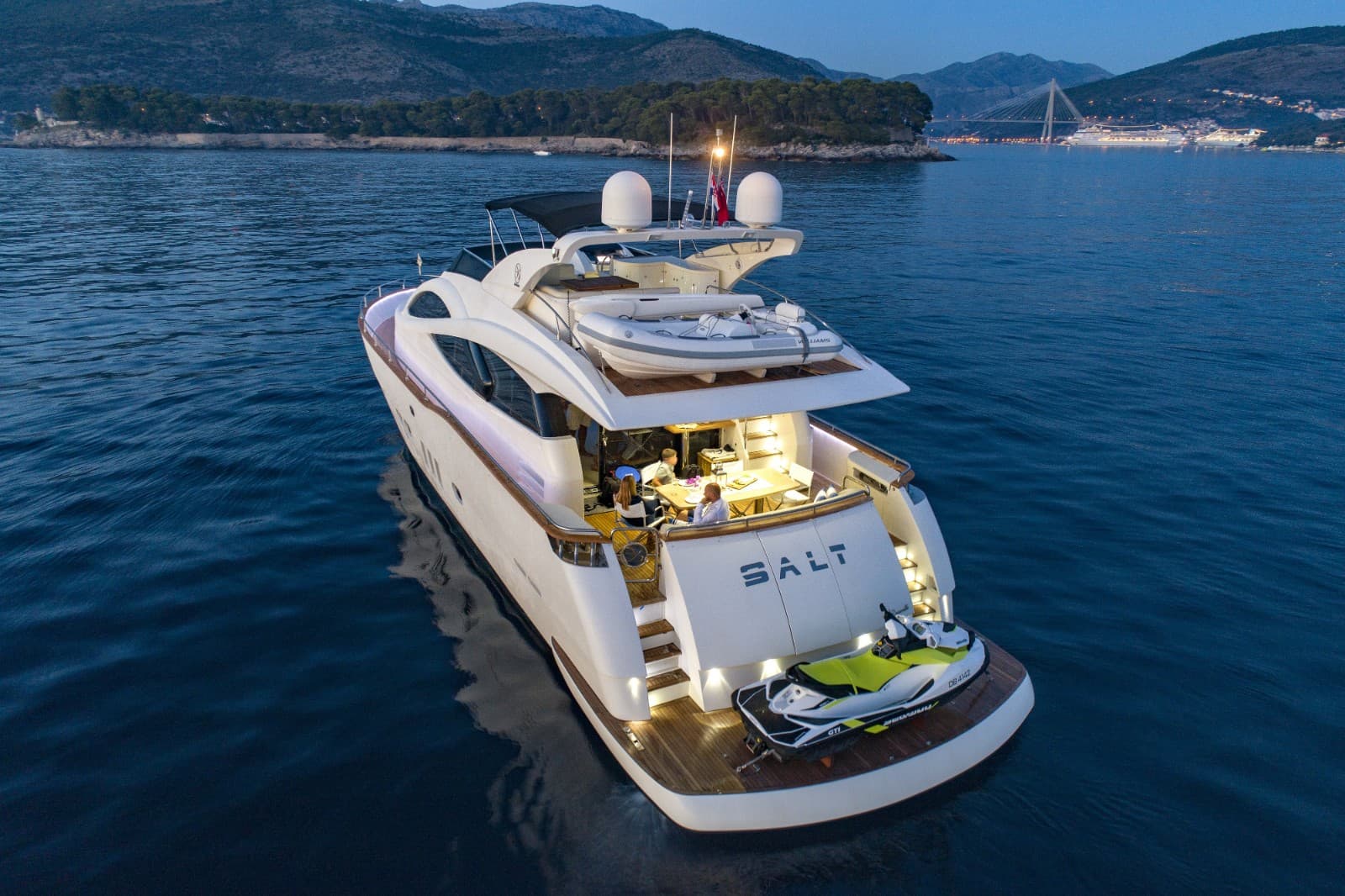 Motor yacht "SALT" with jet ski docked at stern, dining area lit, near mountains at dusk.