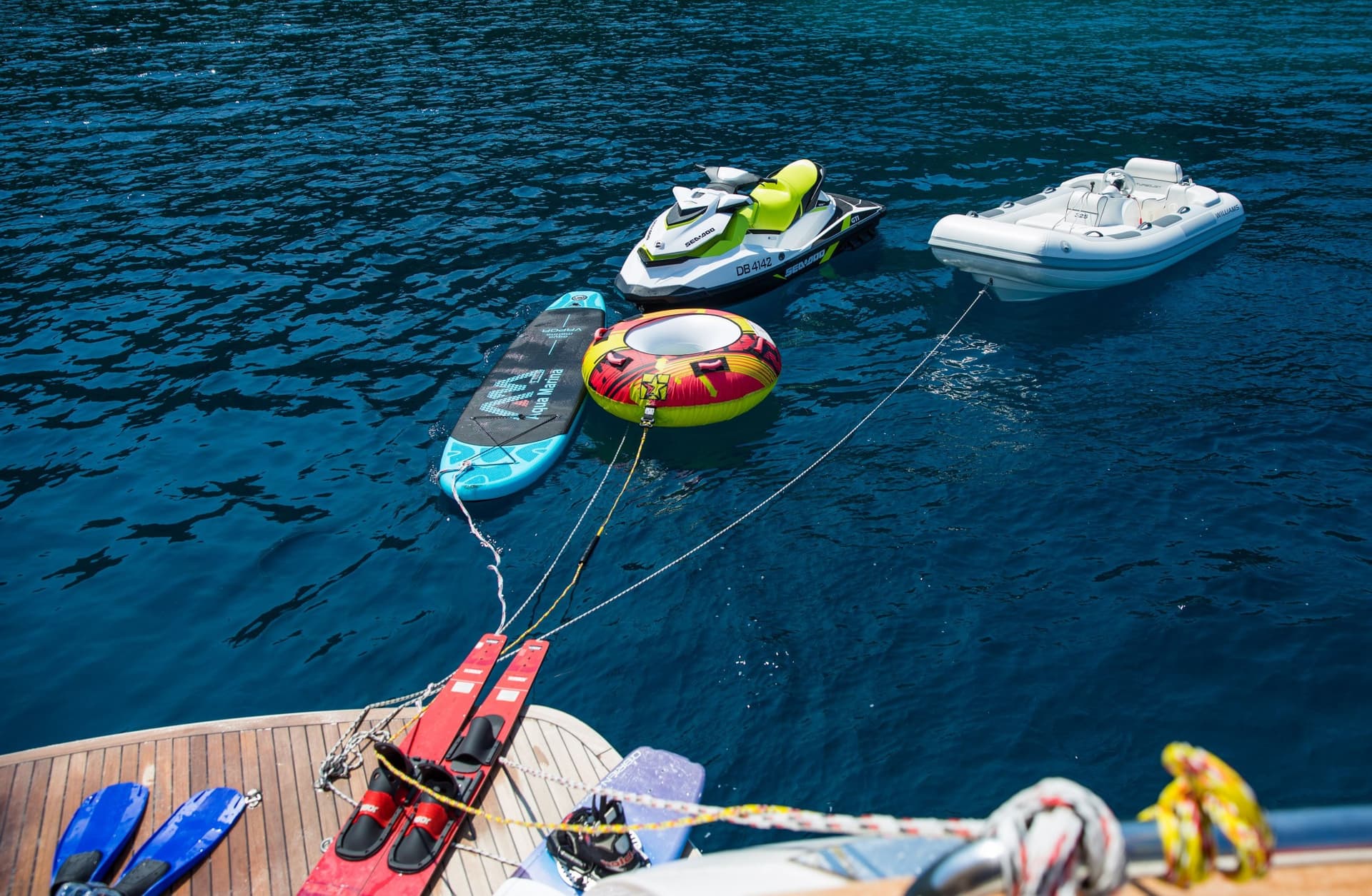 Water toys including jet ski, inflatable, and paddleboard floating near a boat deck with fins and skis.