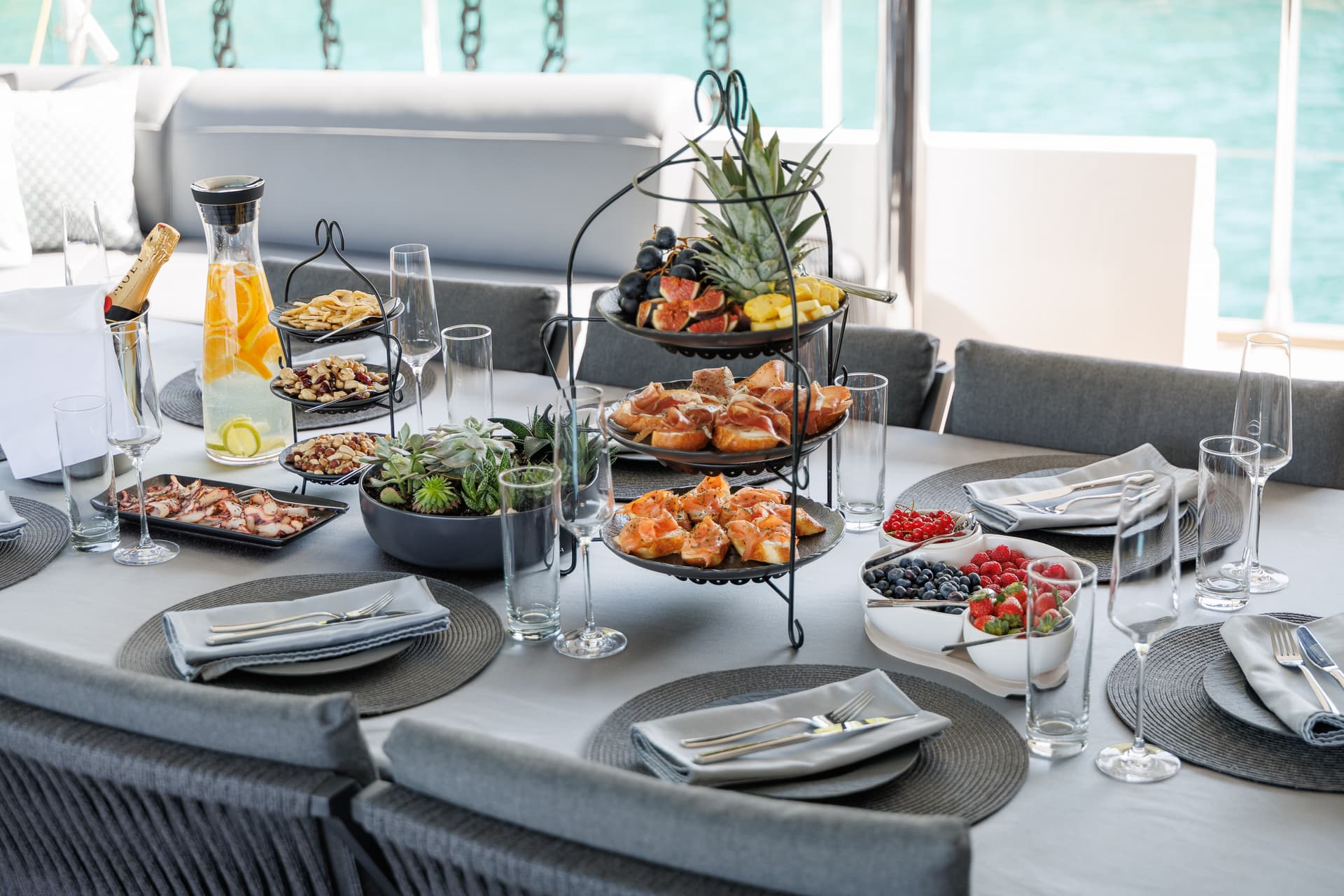 Breakfast spread with fruit, appetizers, and champagne on a yacht deck overlooking turquoise water.