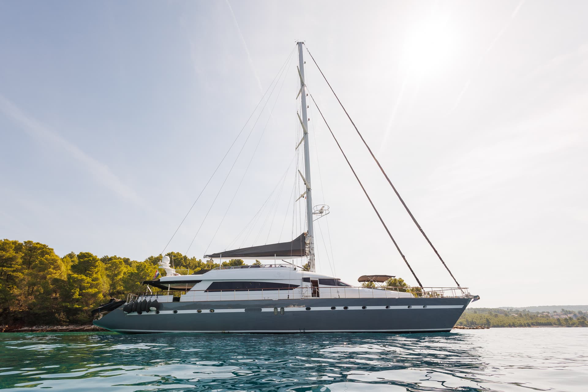 Large sailboat anchored near a lush green coastline under bright sunlight.