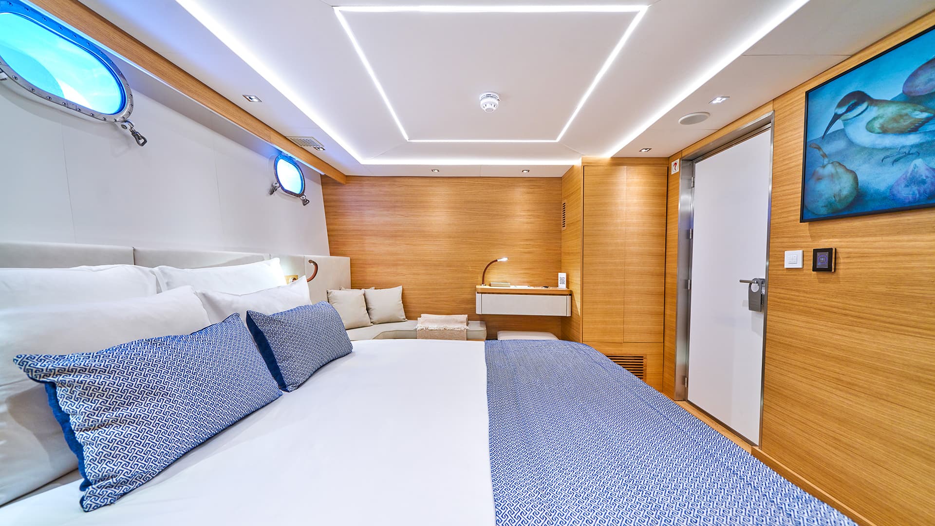 Modern yacht cabin interior with wood paneling, white bedding, blue pillows, and illuminated portholes.