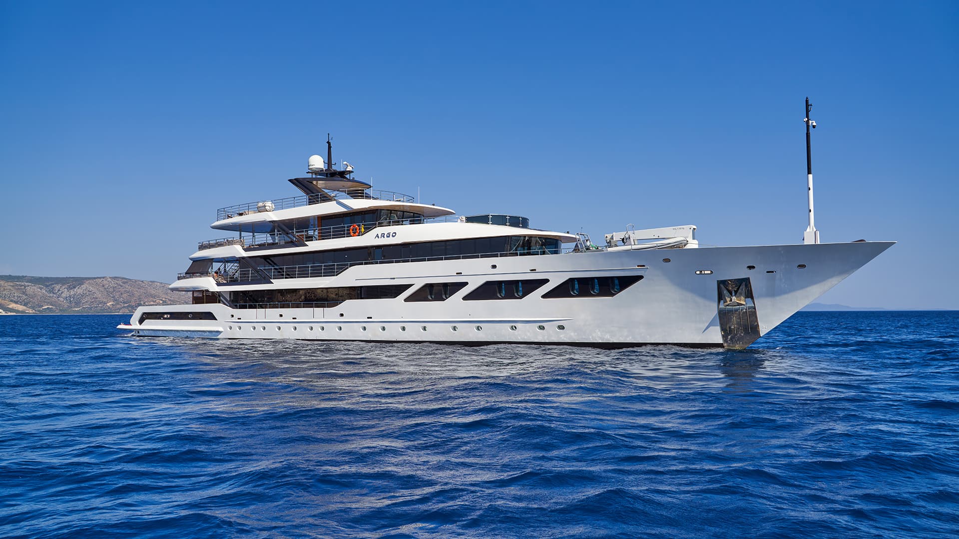 Large white yacht named ARGO cruising on deep blue sea near hilly coastline under clear sky.