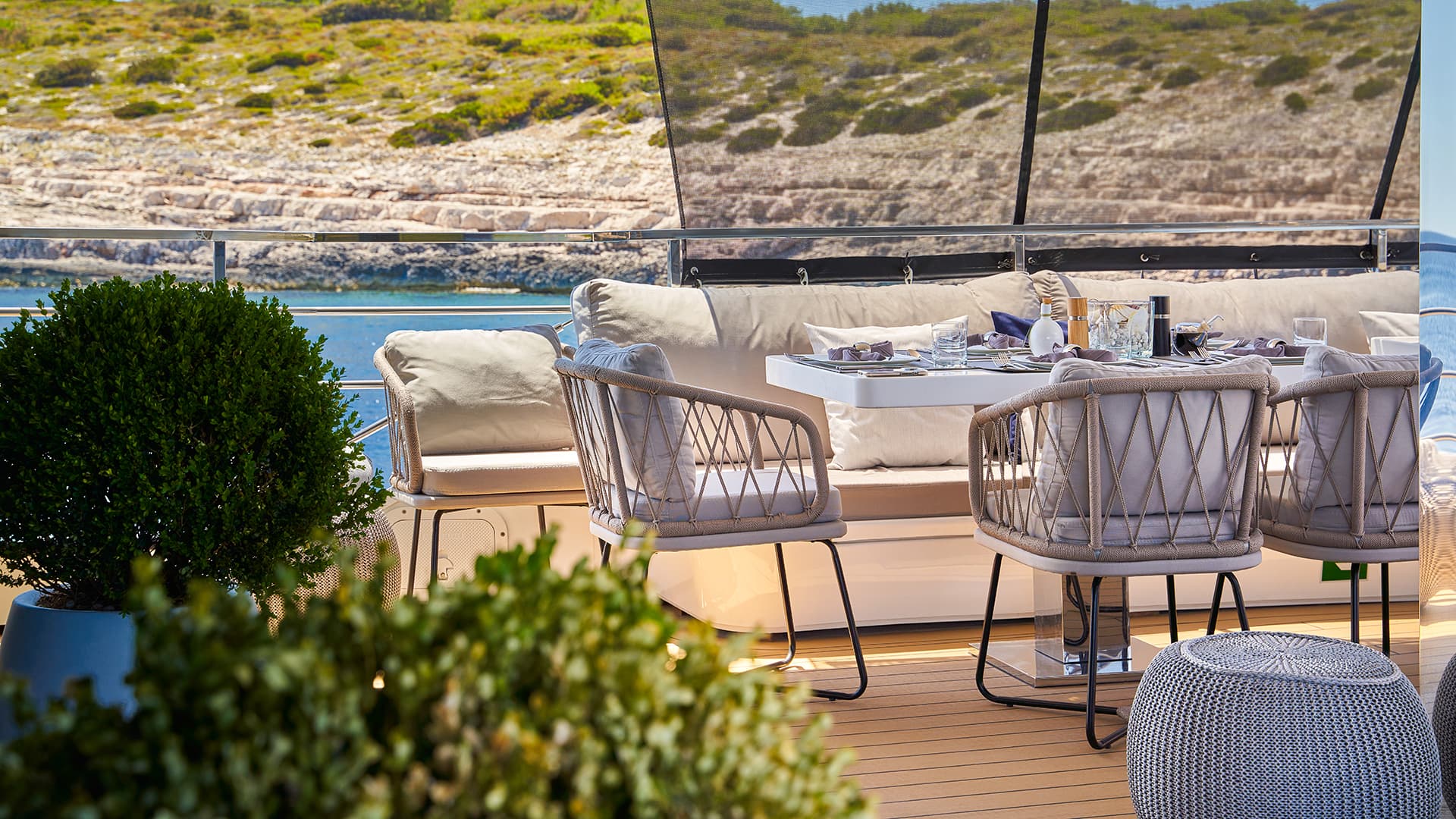 Outdoor dining area on yacht deck with rocky coastline and blue water background