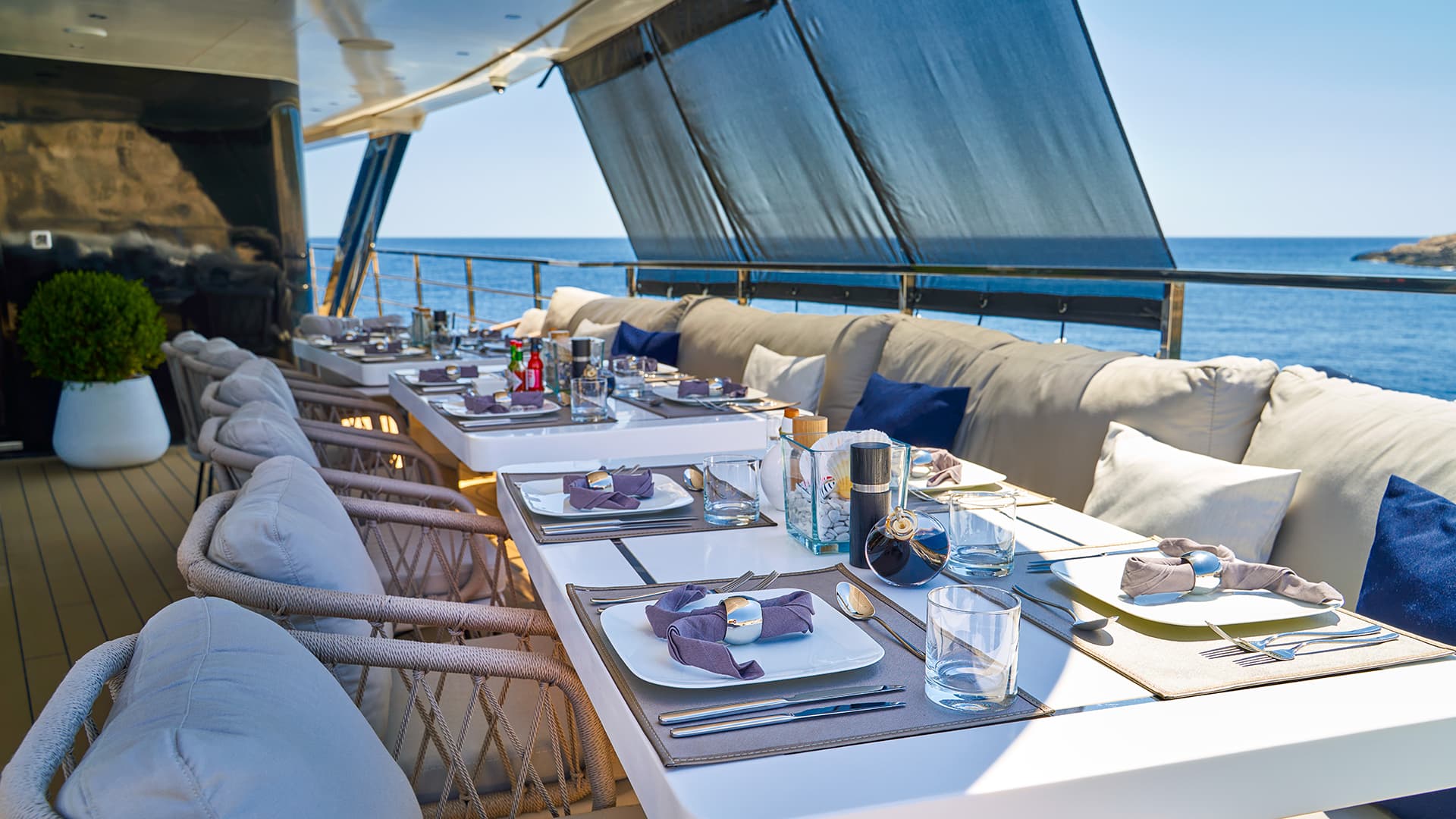 Outdoor dining area set on a yacht deck overlooking the blue sea under awnings.