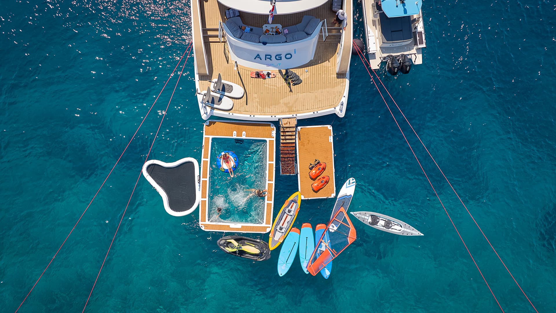 Yacht ARGO with inflatable pool, kayaks, and water toys anchored in clear blue sea.