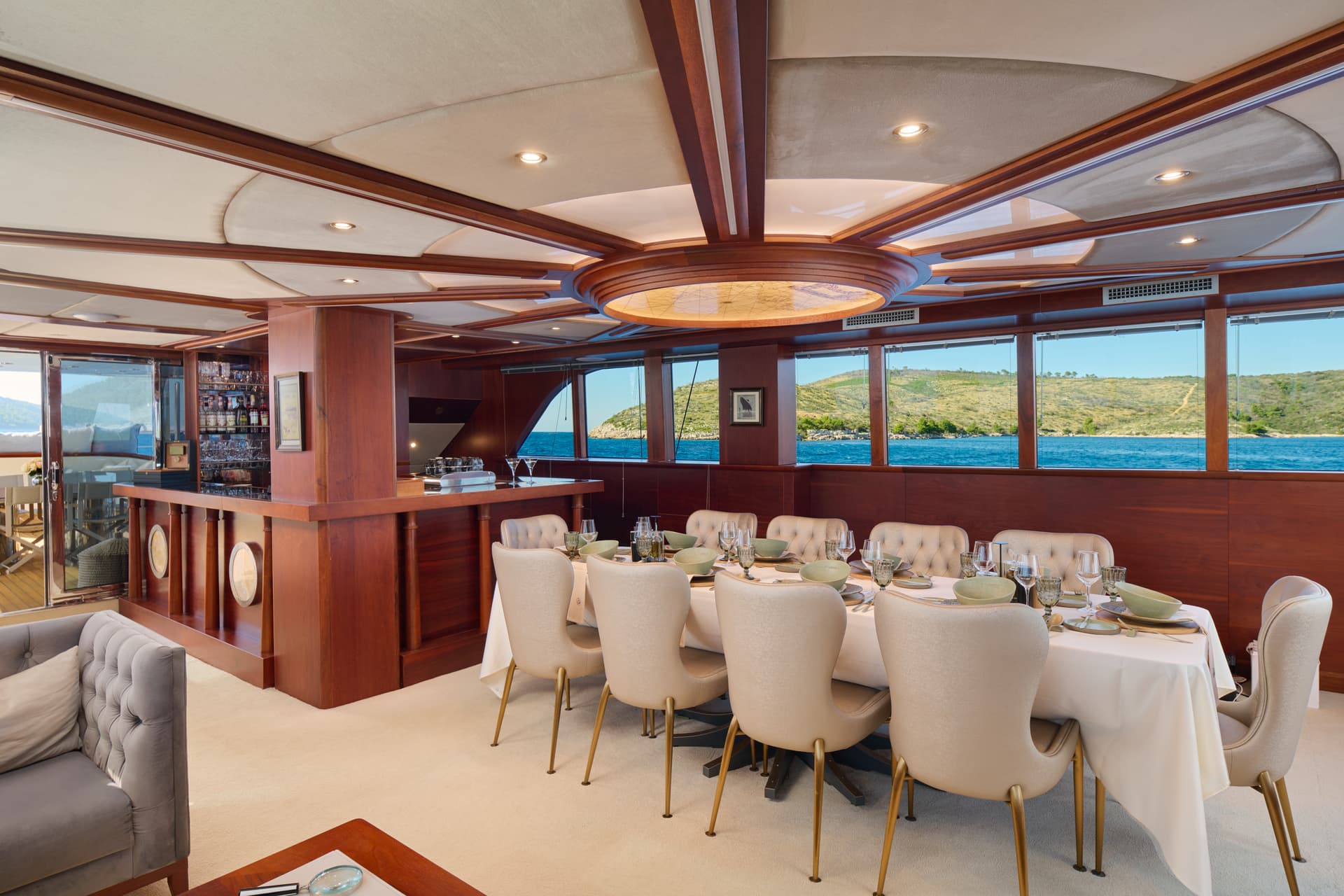 Yacht dining salon with wood paneling, set table, and view of green island coastline.