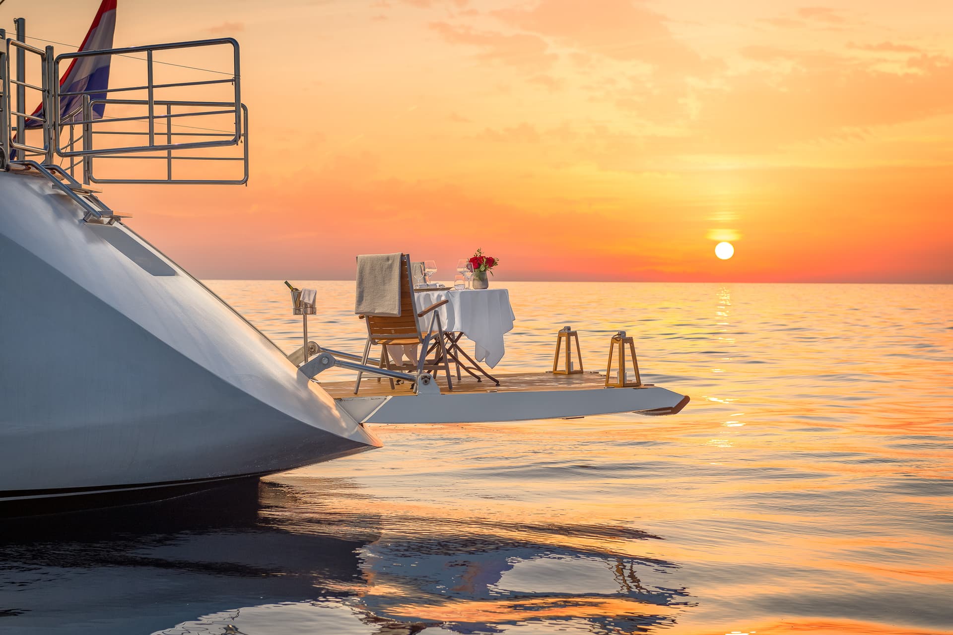 Romantic dinner setup on yacht platform at sunset over calm orange sea water