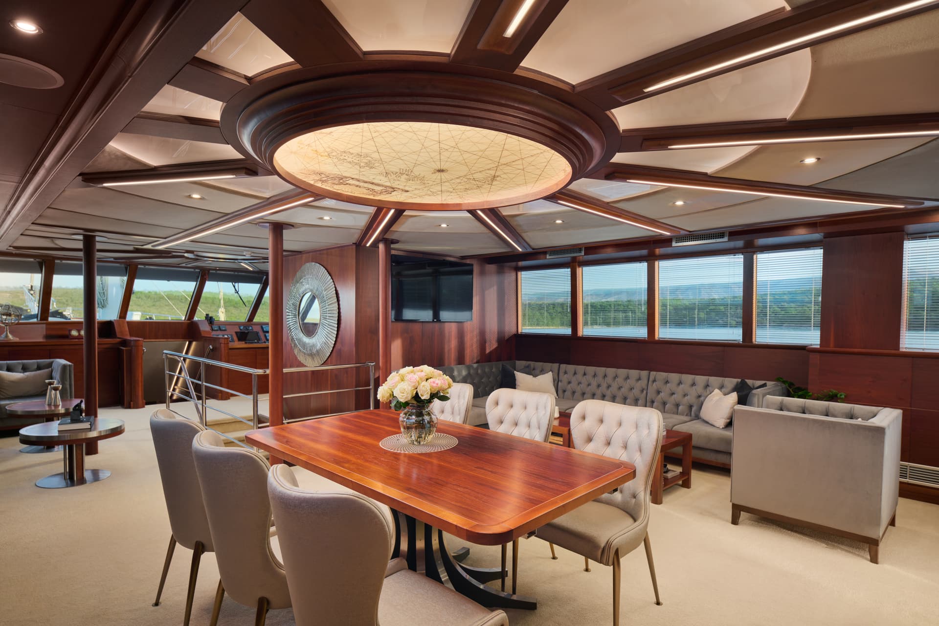 Luxury yacht interior lounge with wood paneling, dining table, and view of green coastline.