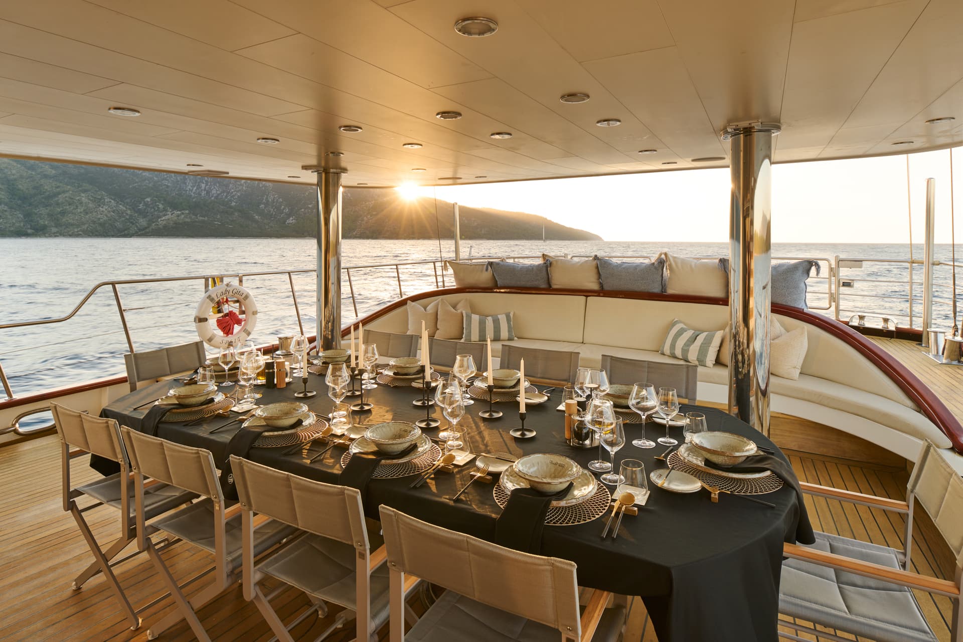 Outdoor dining table set on a yacht deck at sunset near a coastline.