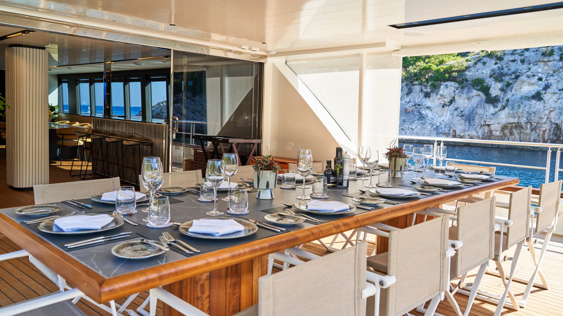 Outdoor dining table set on a yacht deck next to a rocky, vegetated coastline.