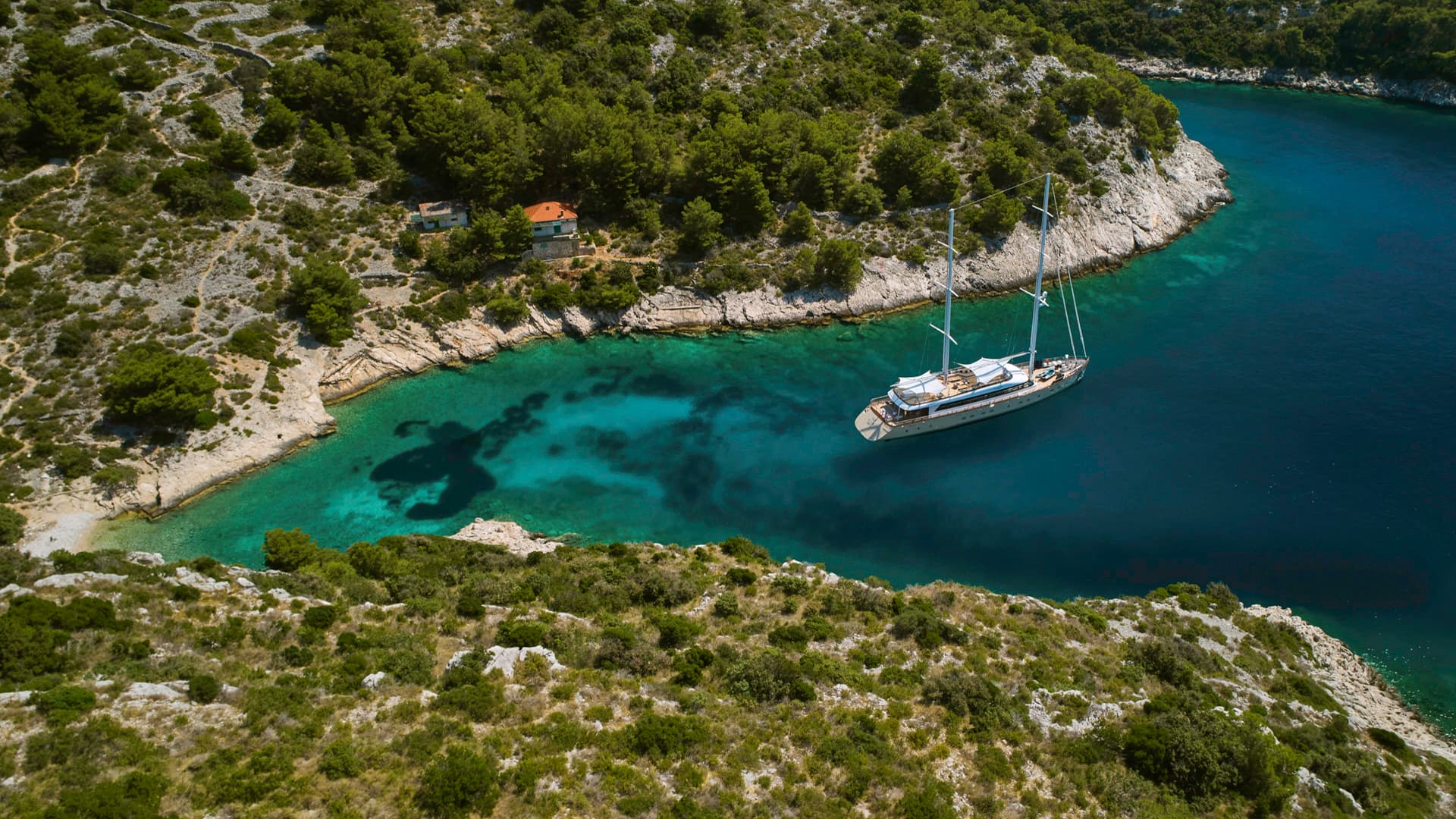 Large sailboat anchored in turquoise cove surrounded by green, rocky coastline.