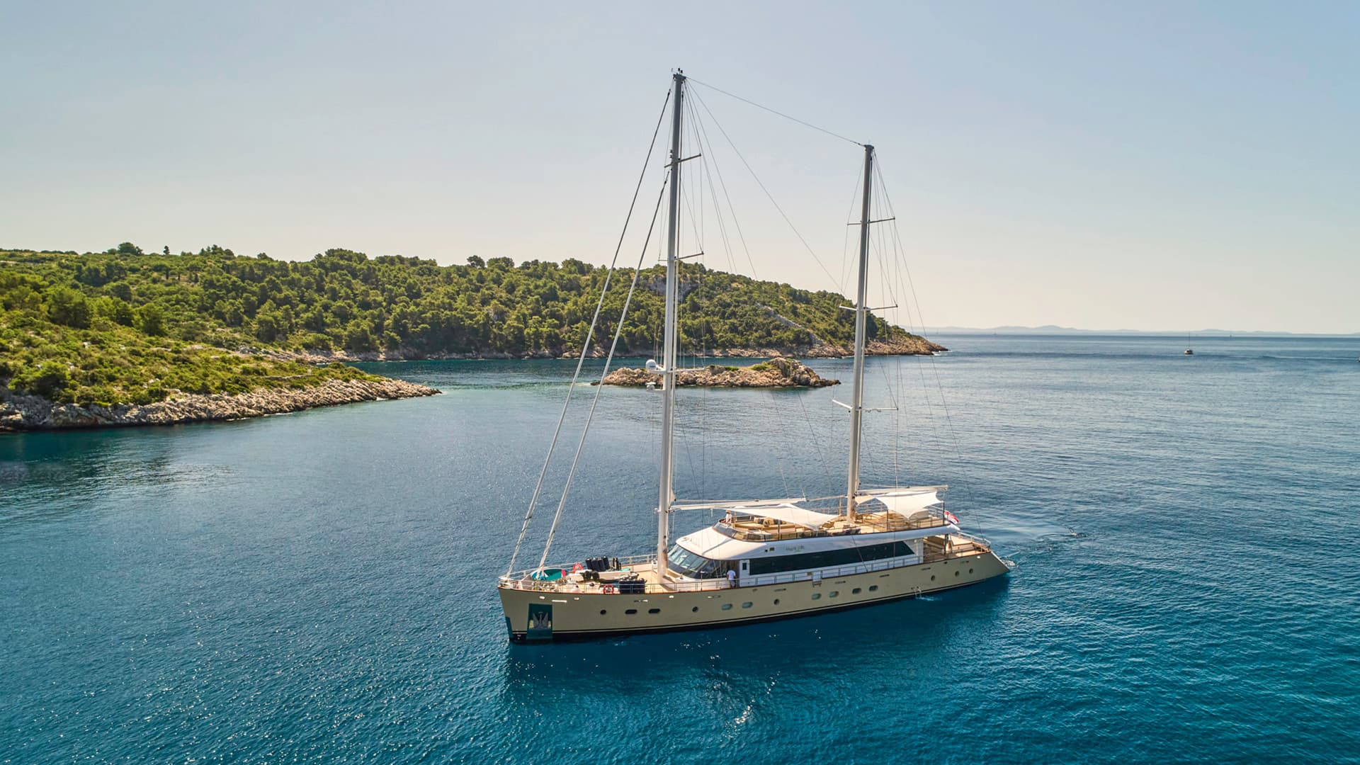 Large sailboat anchored in turquoise cove near densely wooded coastline under clear sky