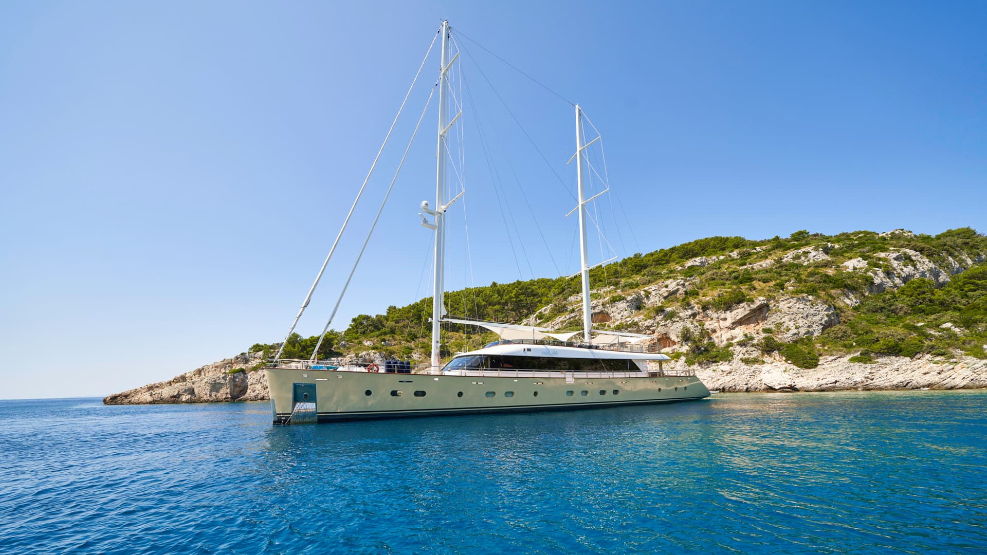 Large sailing yacht anchored near rocky, green coastline under clear blue sky