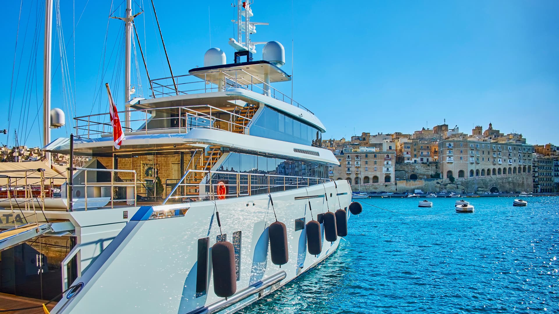 Large white yacht moored in bright blue water with historic waterfront buildings ashore.