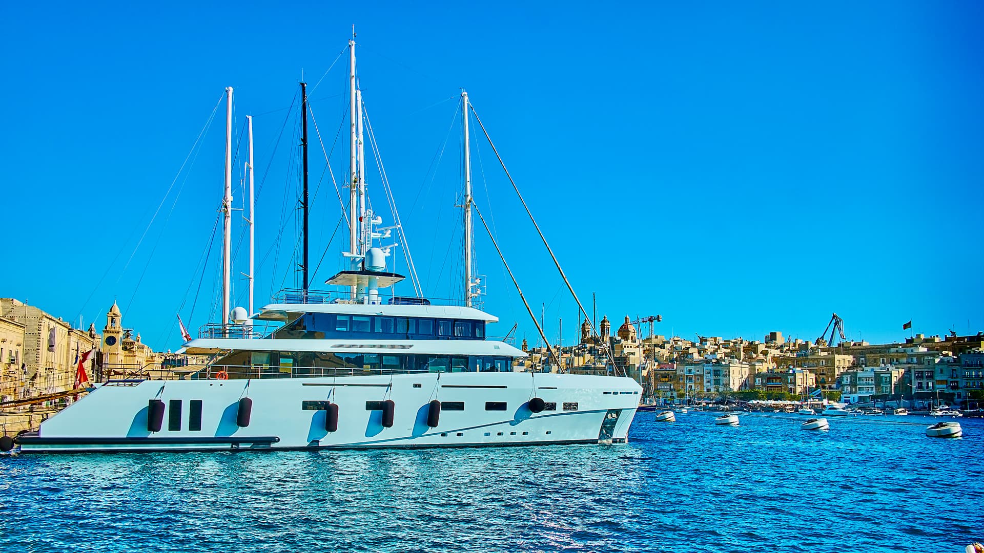 Large white yacht moored in blue harbor water near historic city skyline under bright blue sky.