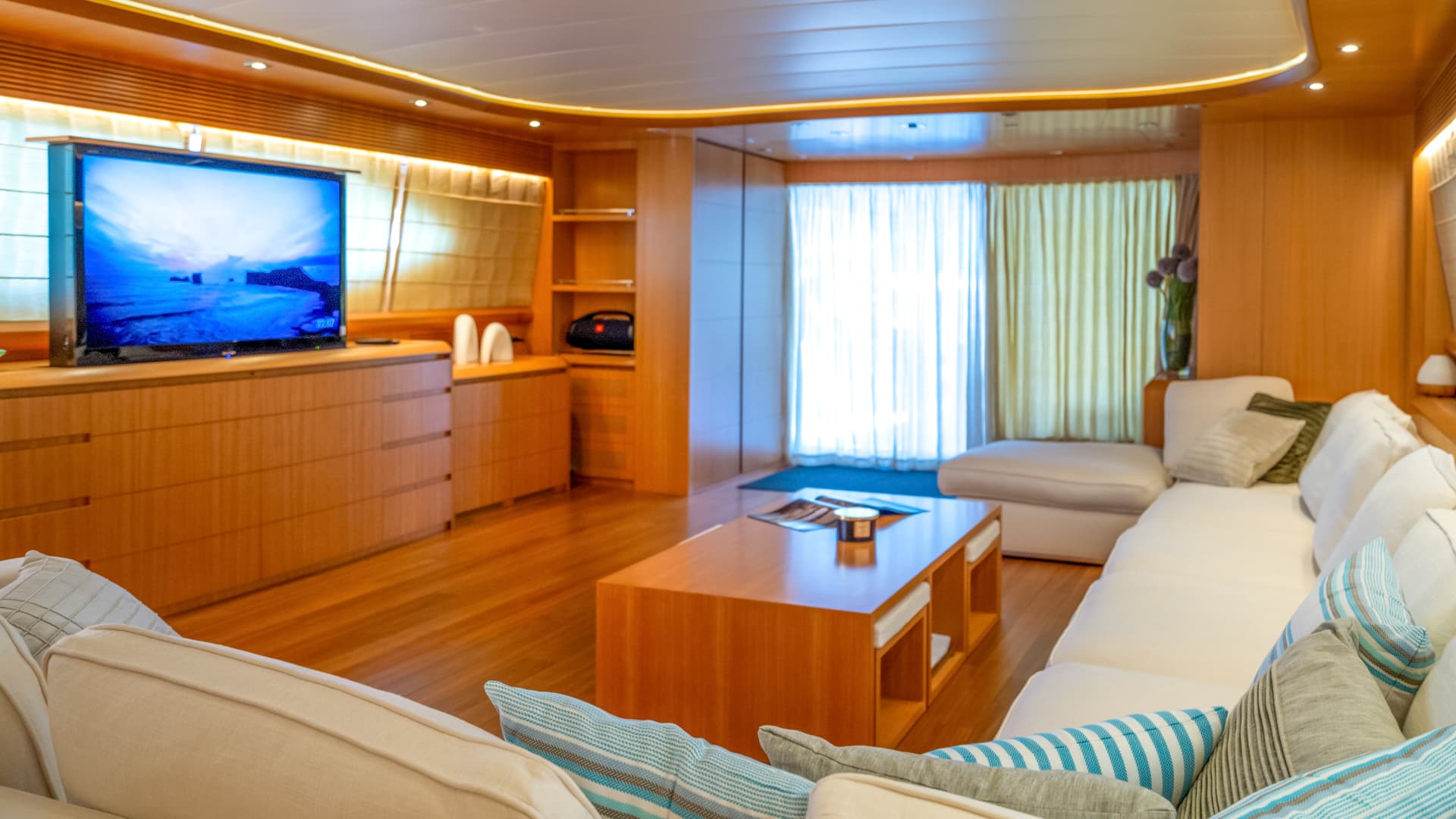 Luxurious yacht interior with wood paneling, white sectional sofa, and large television displaying a blue seascape.