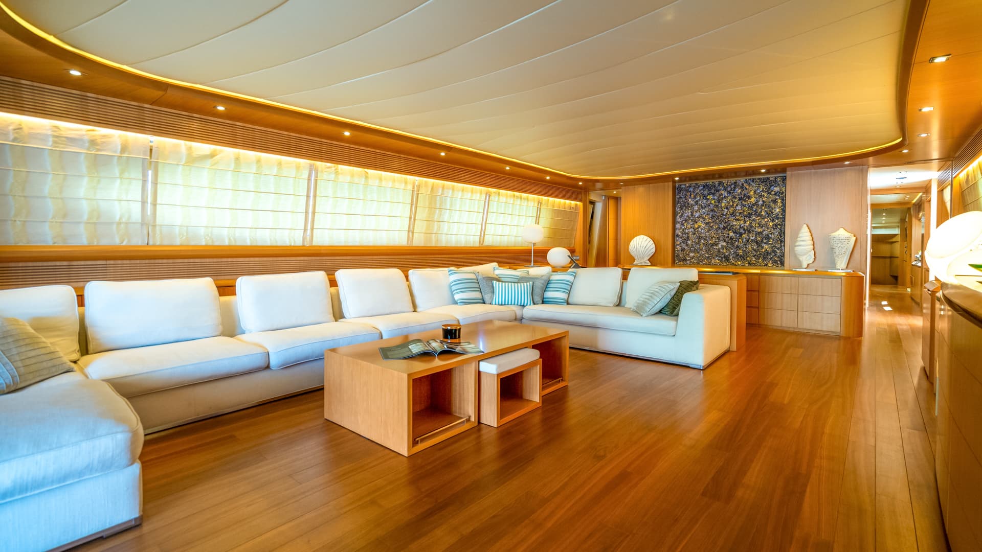 Luxurious yacht salon with white sectional sofa, wood paneling, and large windows with blinds.