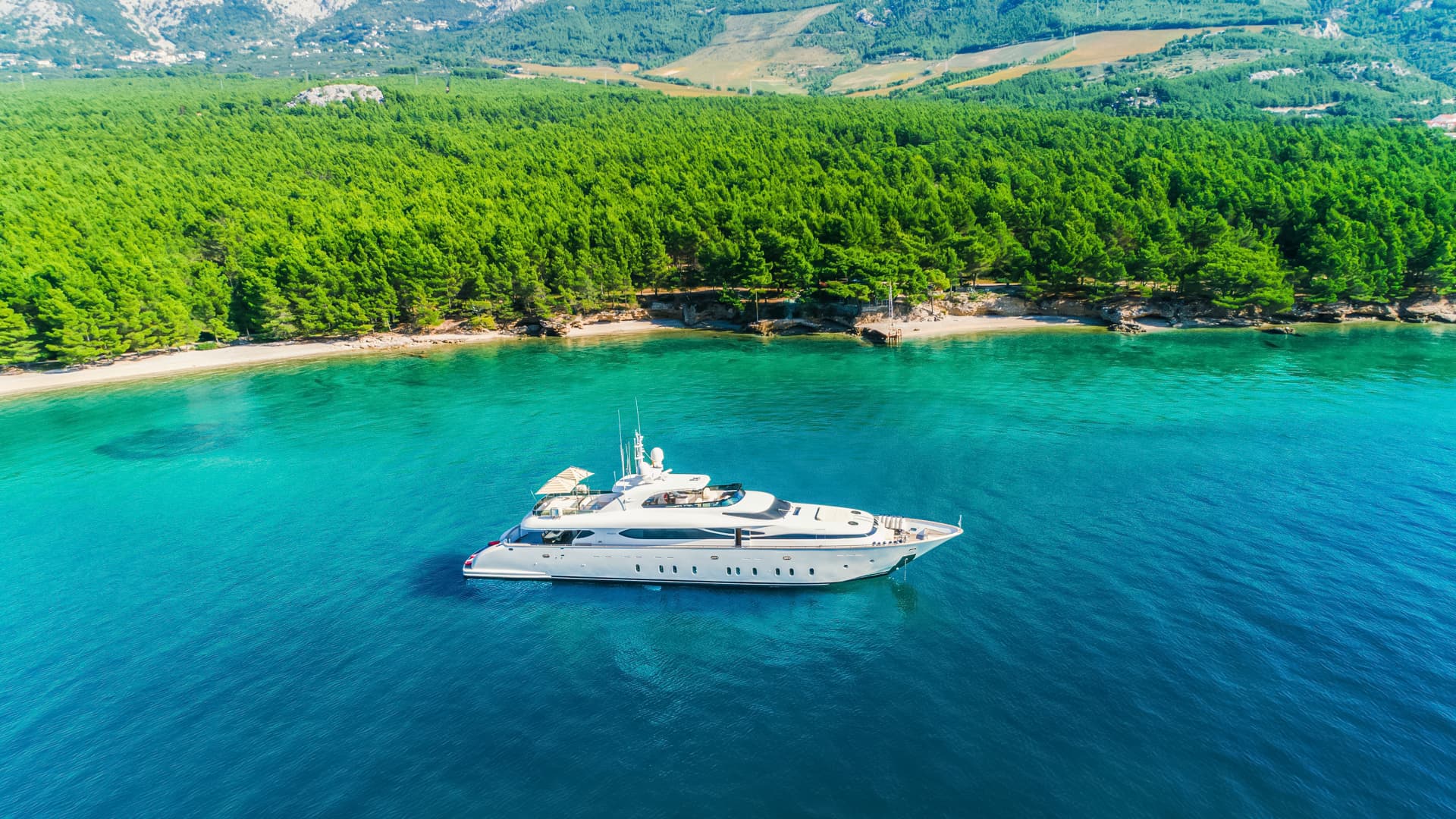 Luxury yacht anchored in turquoise water near a forested coastline and beach.