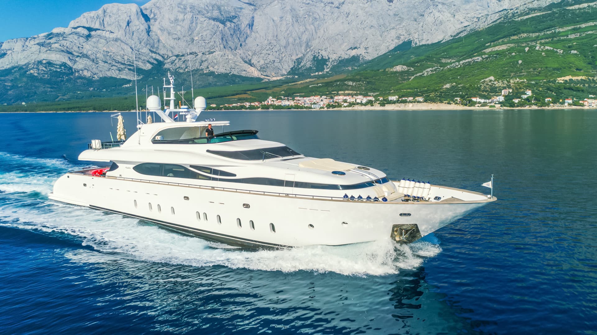 White motor yacht cruising on blue water with steep, rocky mountains and coastal town backdrop.