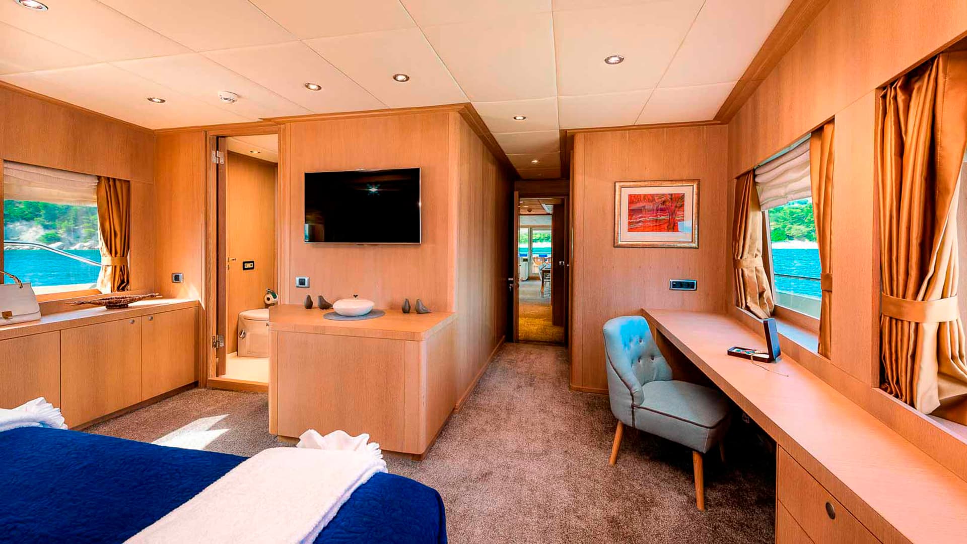 Master cabin interior with wood paneling, blue bedding, and view of turquoise water.