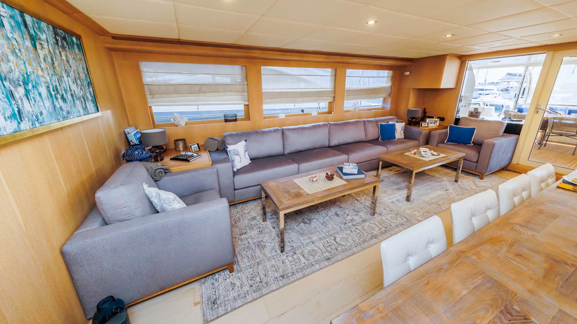 Main salon of a yacht with wood paneling, gray sectional sofa, and dining area.