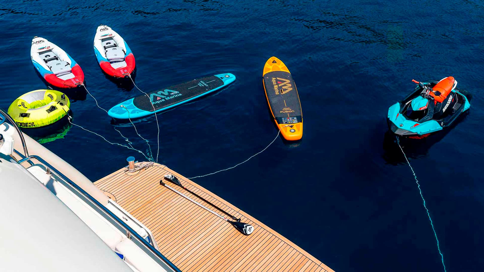 Water toys including paddleboards, inflatable kayaks, and a jet ski tethered to a yacht's teak deck.