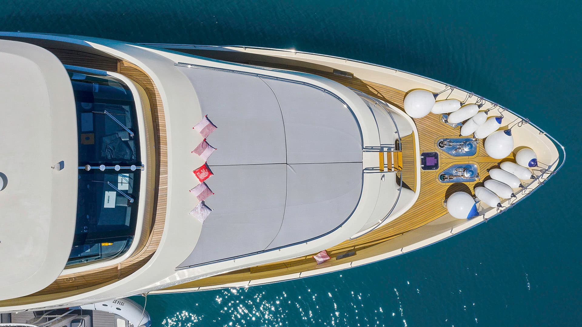 Aerial view of a luxury yacht foredeck with white fenders and decorative pillows on turquoise water.
