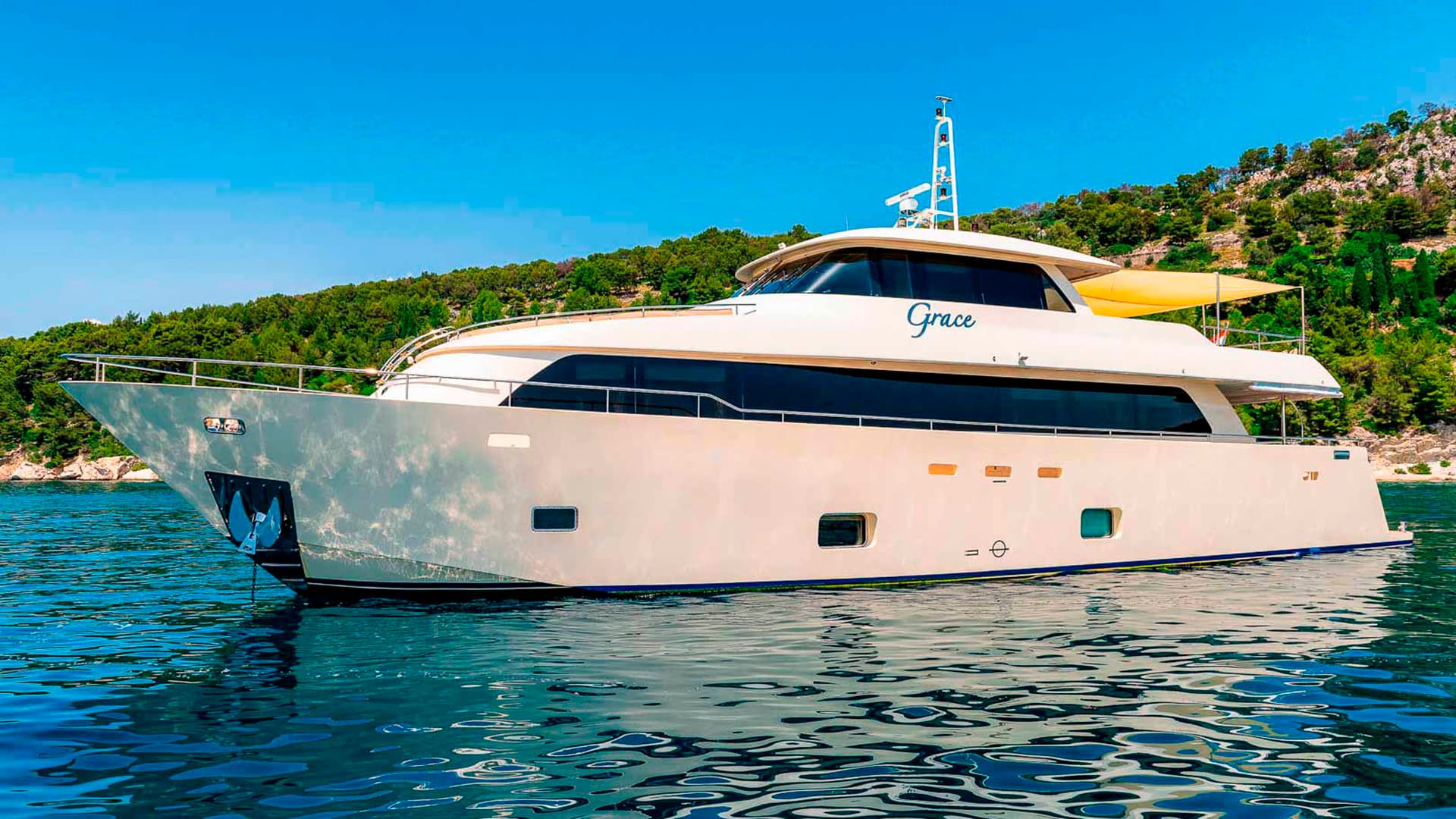 Motor yacht 'Grace' anchored near a lush, green, rocky coastline under a clear blue sky.