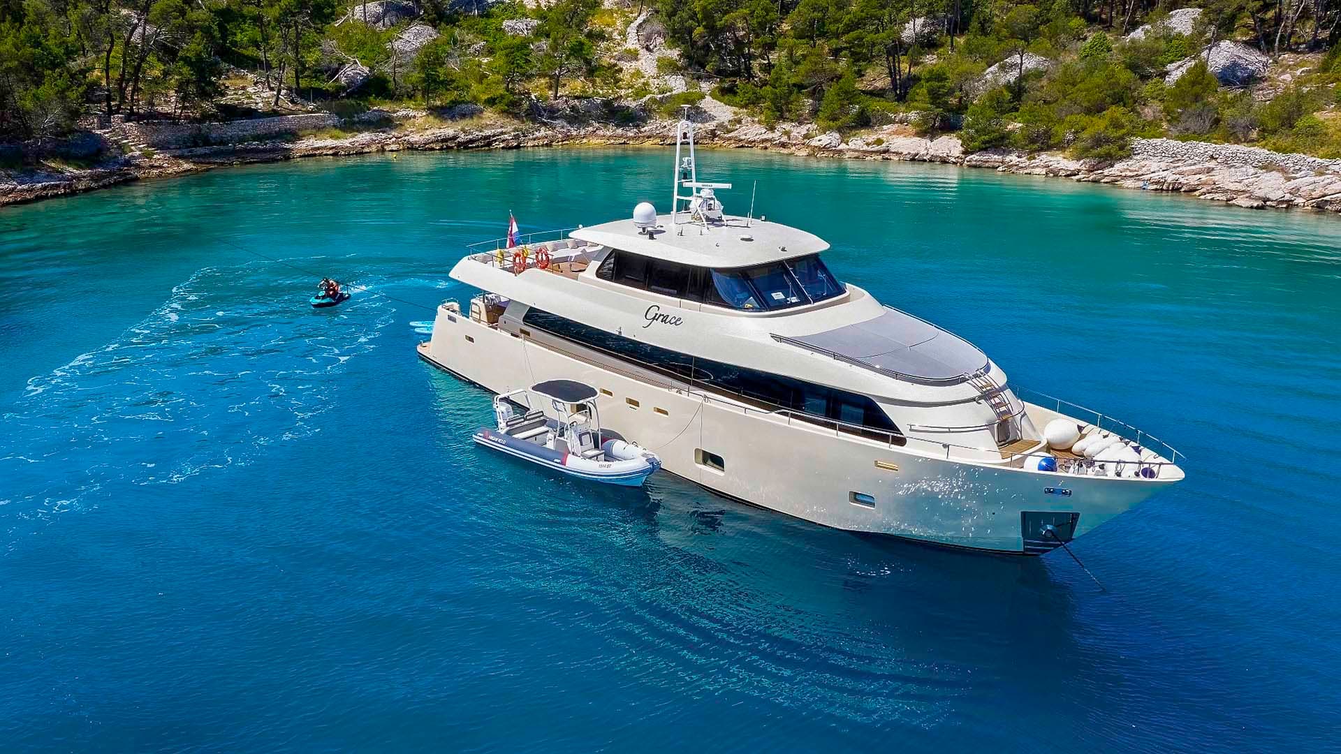 Large yacht named Grace anchored in turquoise cove with wooded rocky shoreline; jet ski activity nearby.