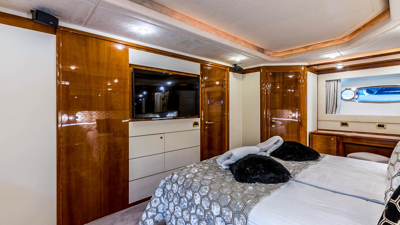 Master cabin interior with glossy wood, flat-screen TV, and porthole window view of blue water.