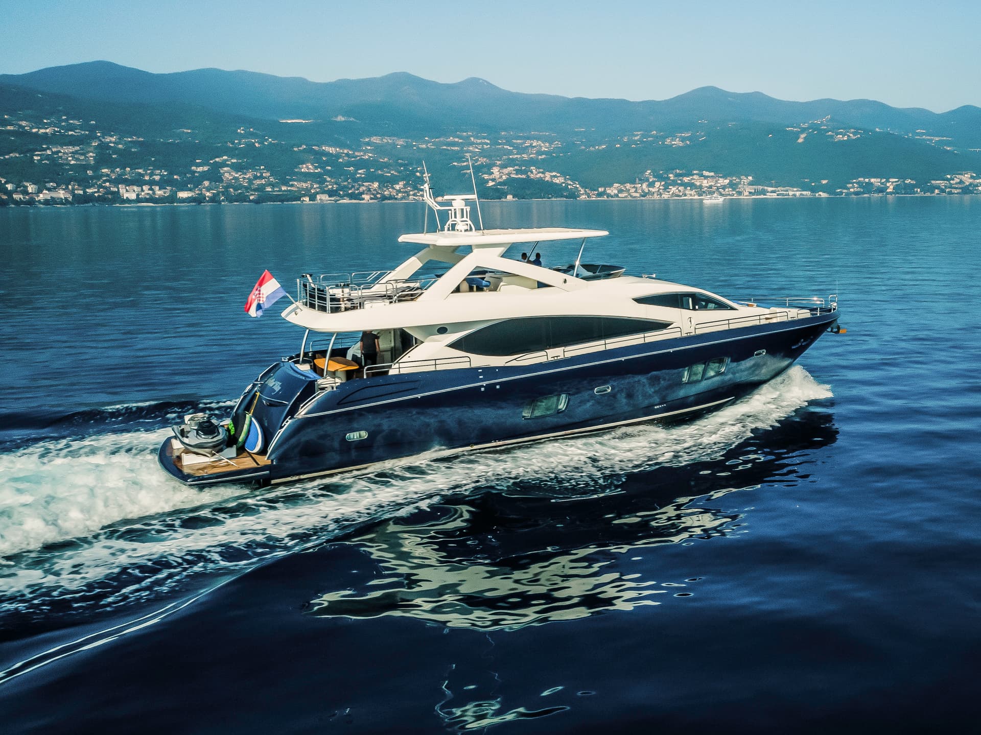 Luxury yacht speeding on dark blue water with Croatian flag near mountainous coastline.
