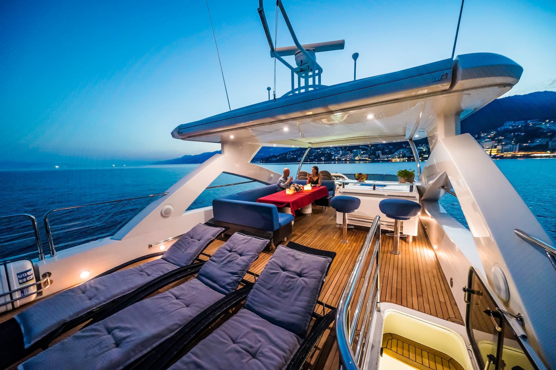 Yacht deck with sun loungers and couple dining at dusk near illuminated Croatian coastline.