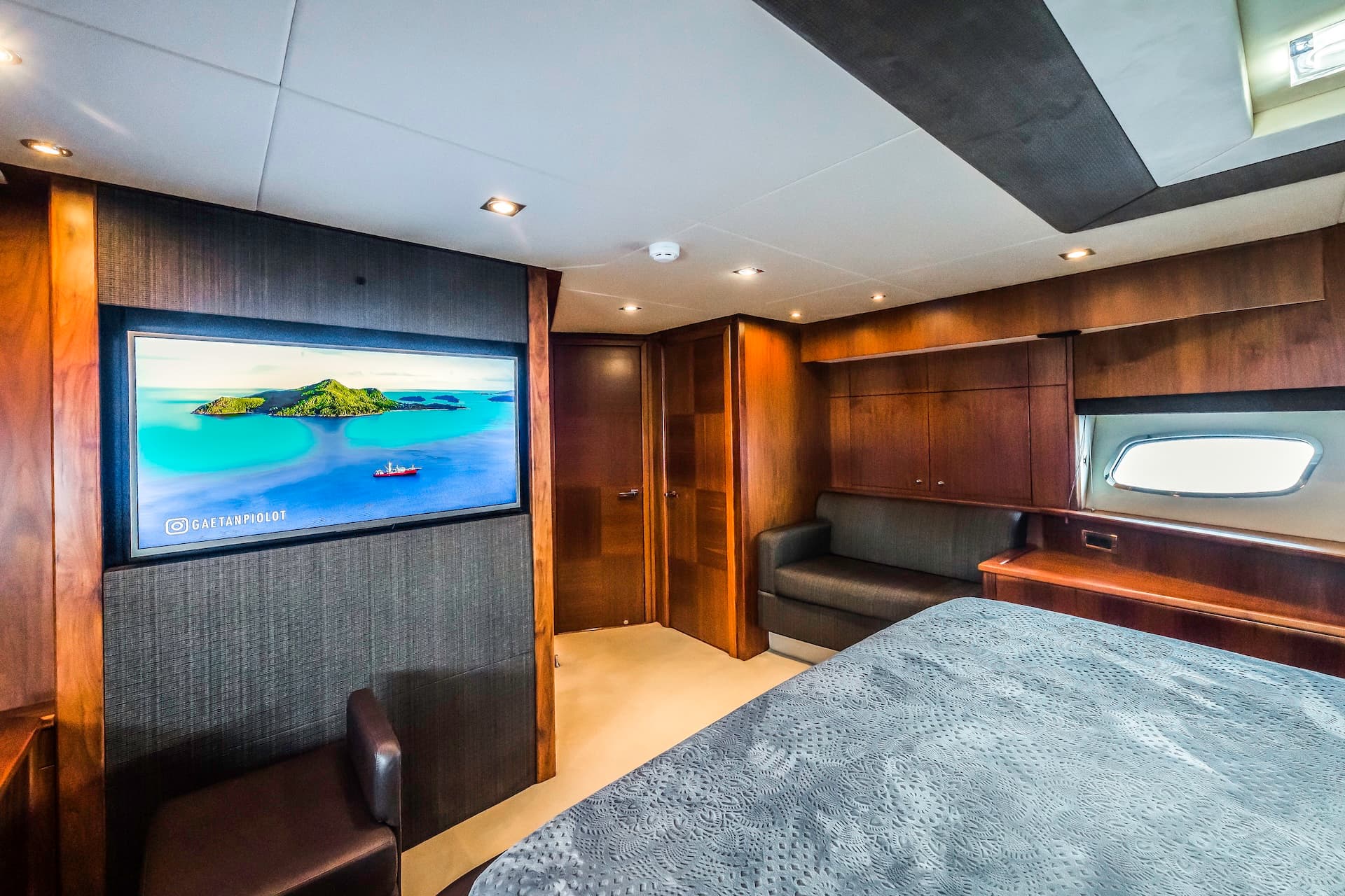Yacht cabin interior with wood paneling, a large TV displaying a tropical island, and a bed.