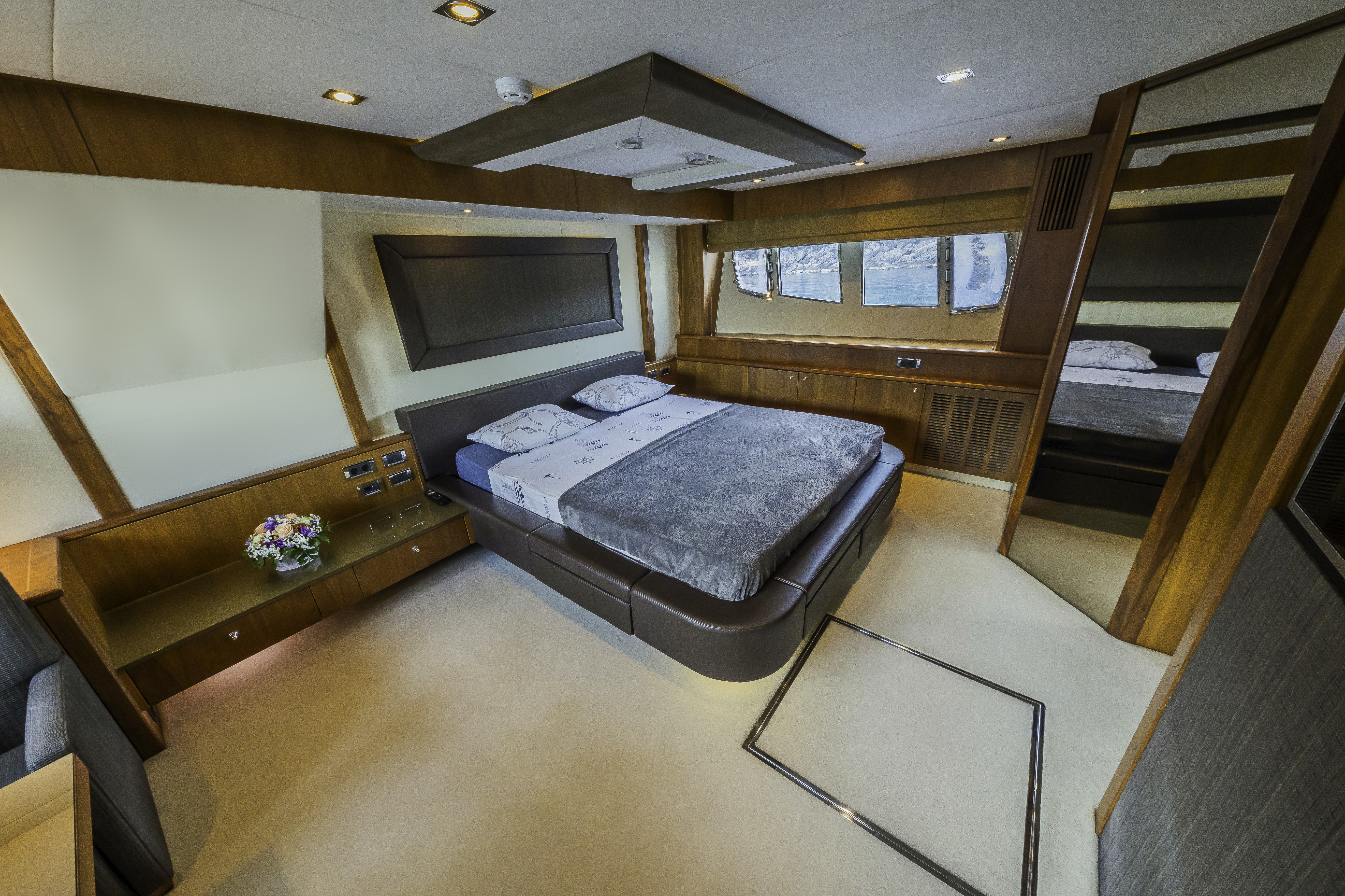Yacht cabin bedroom with wood trim, view of rocky coastline and blue water through windows.